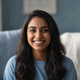 Headshot portrait of a happy young adult South Asian woman with a genuine smile, wearing a blue top, against a soft blue and 